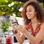 Shot of beautiful glad young female with Afro hairdo, types number of credit card on smart phone, makes purchase online or checks bank account, recreats in outdoor cozy cafe with fresh cocktail
